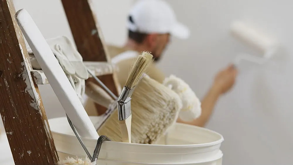 Maler streicht eine weisse Wand mit der Rolle, im Vordergrund Pinsel und Farbeimer auf der Leiter